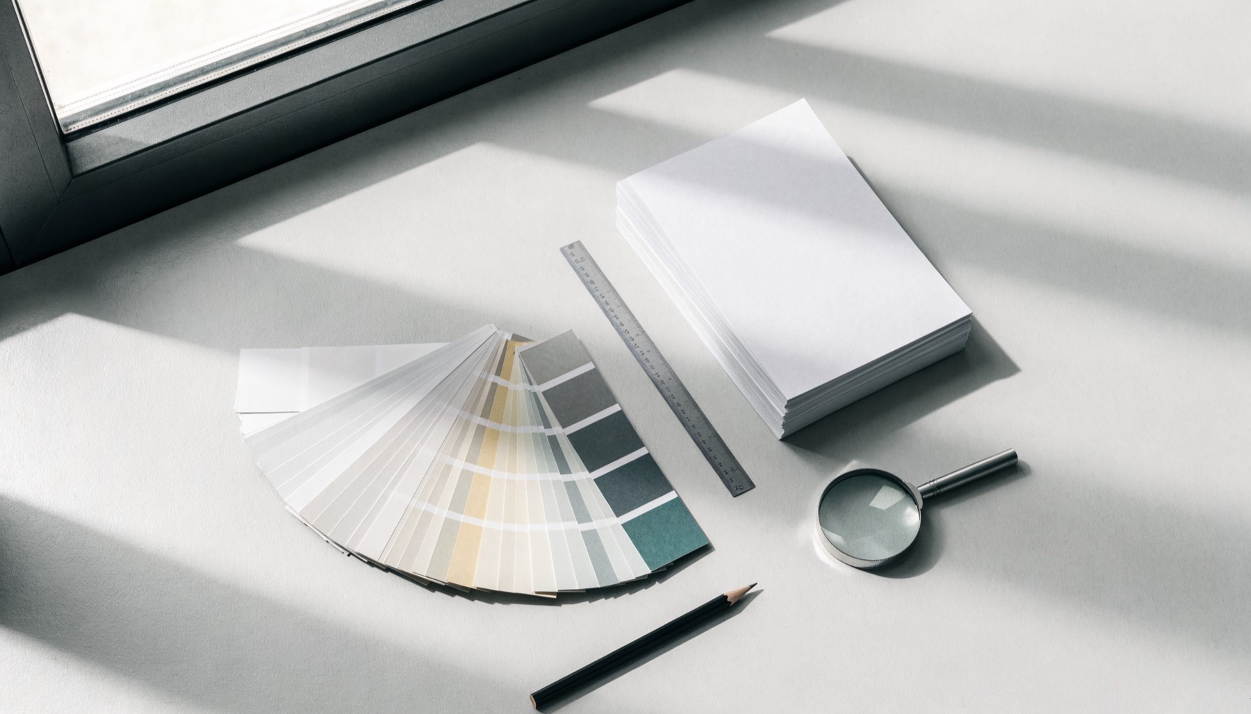 A fan of colour swatch chips, a stack of blank proof sheets, a steel ruler, a pencil and a loupe on a designer's desk