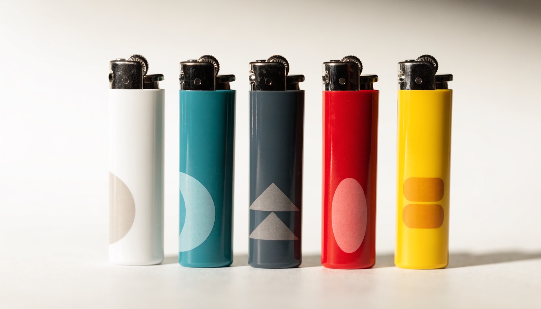 Five promotional lighters in white, teal, charcoal, red and yellow, each decorated with a simple abstract mark inside the imprint area